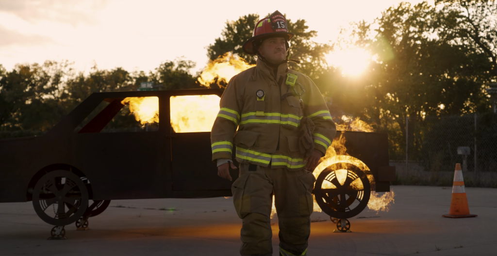 Firefighter in uniform walking with burning car in background