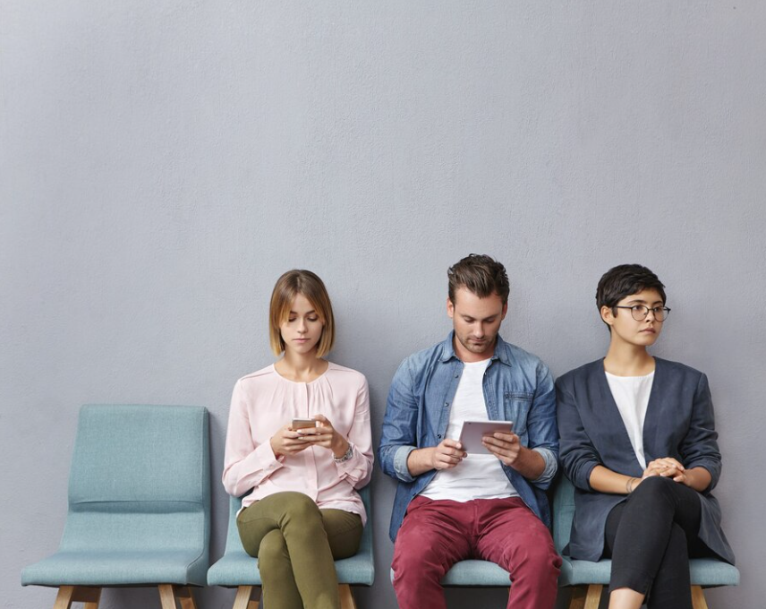 Three individuals sit on chairs against a blue wall, engrossed in their devices