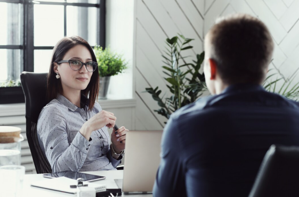 A woman in glasses converses with a man sitting in front of her in an office setting