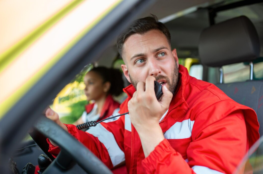 man paramedic talking by portable radio while sitting in ambulance, woman behind in the background 